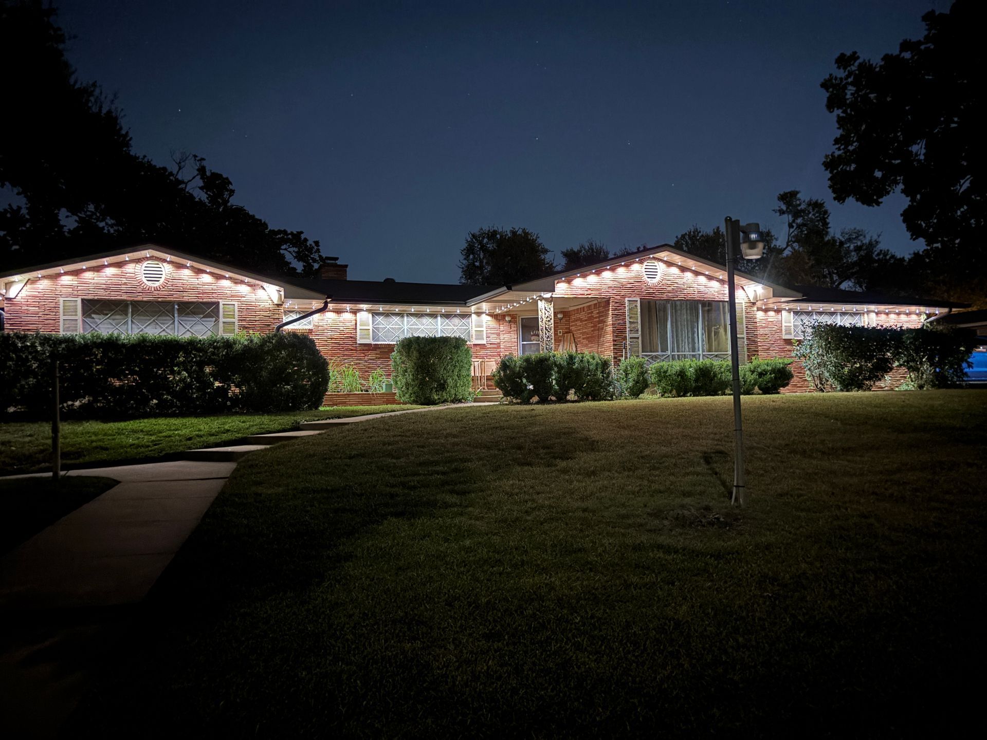 A large house outlined with white Christmas lights at night, with a dark lawn and trees in the foreground.