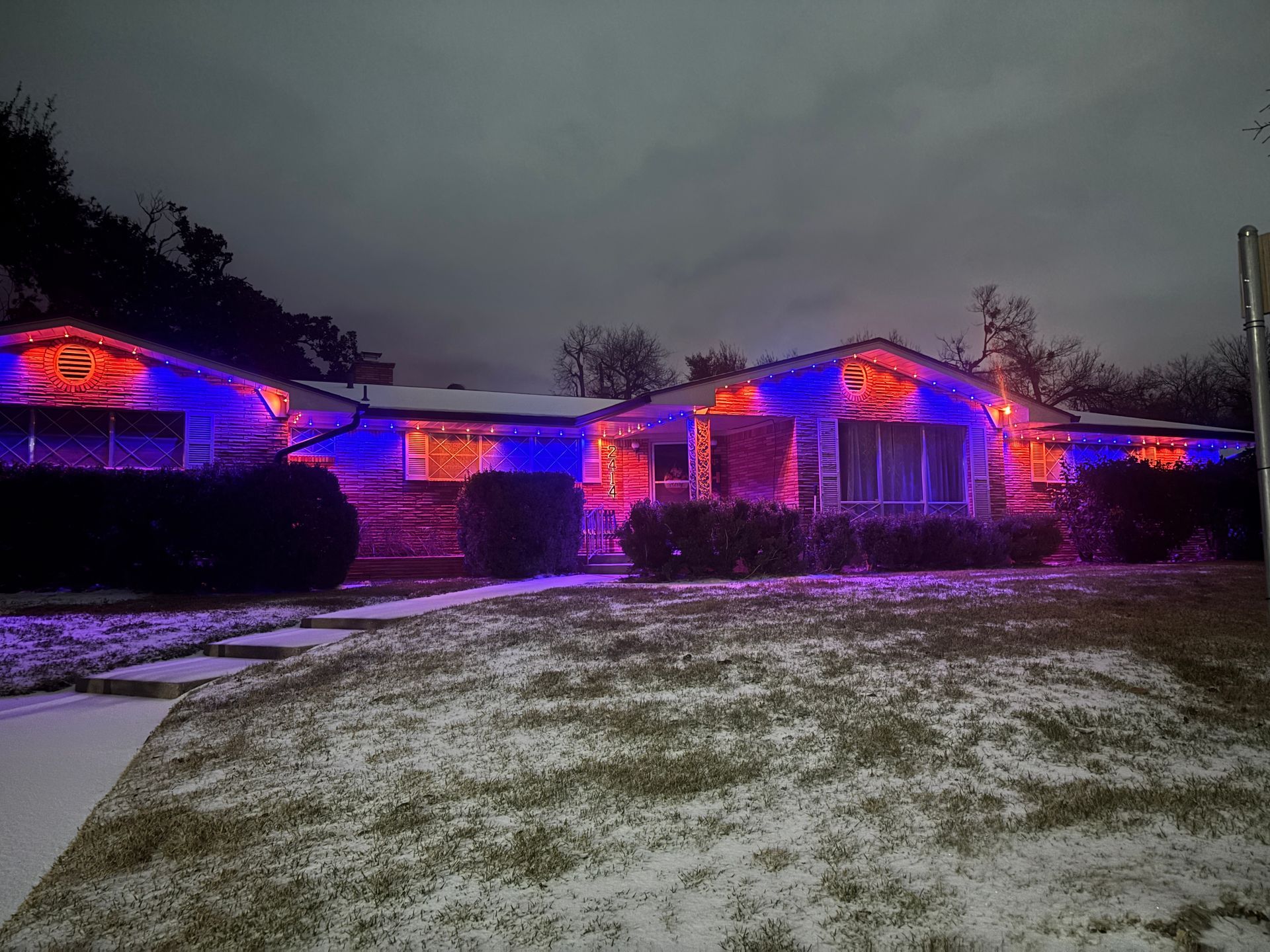 Single-story house at night with purple and red holiday lights in a snowy yard