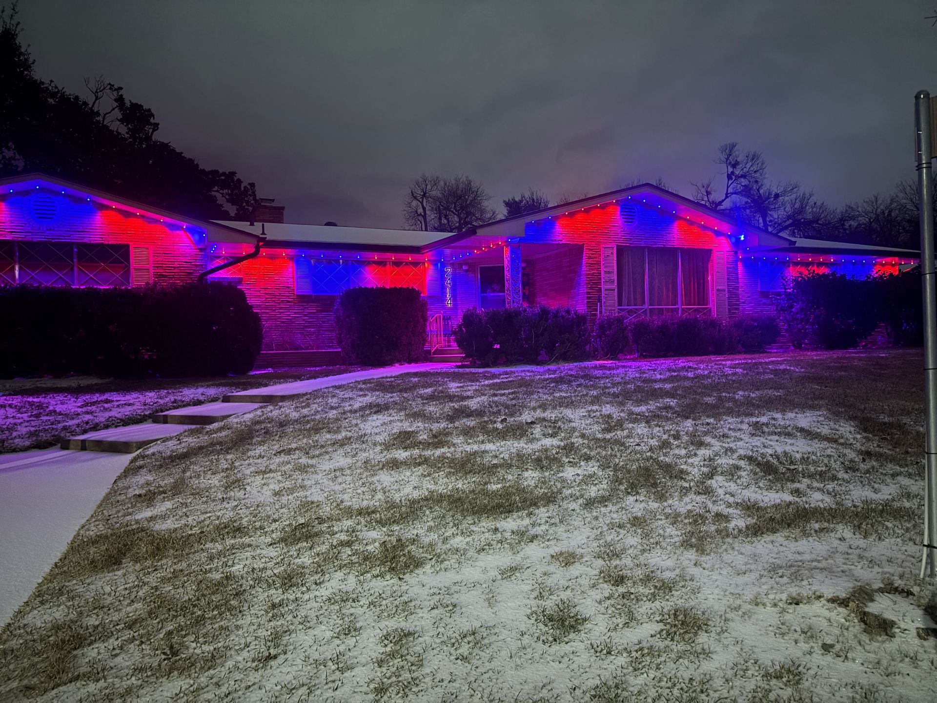 House lit with red and blue holiday lights at dusk, with a light dusting of snow on the lawn.