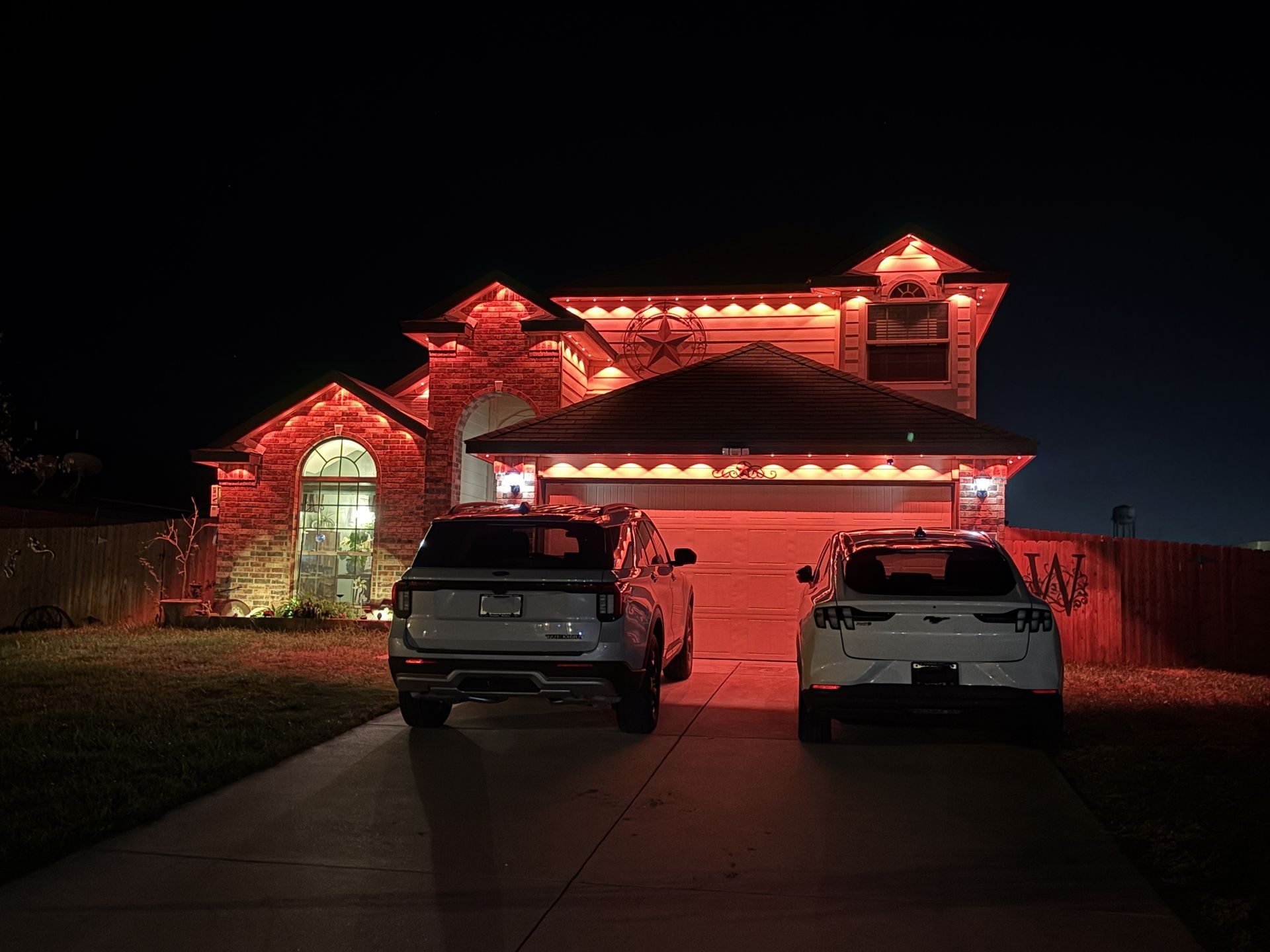 House lit with red-orange holiday lights at night, with two SUVs parked in the driveway.