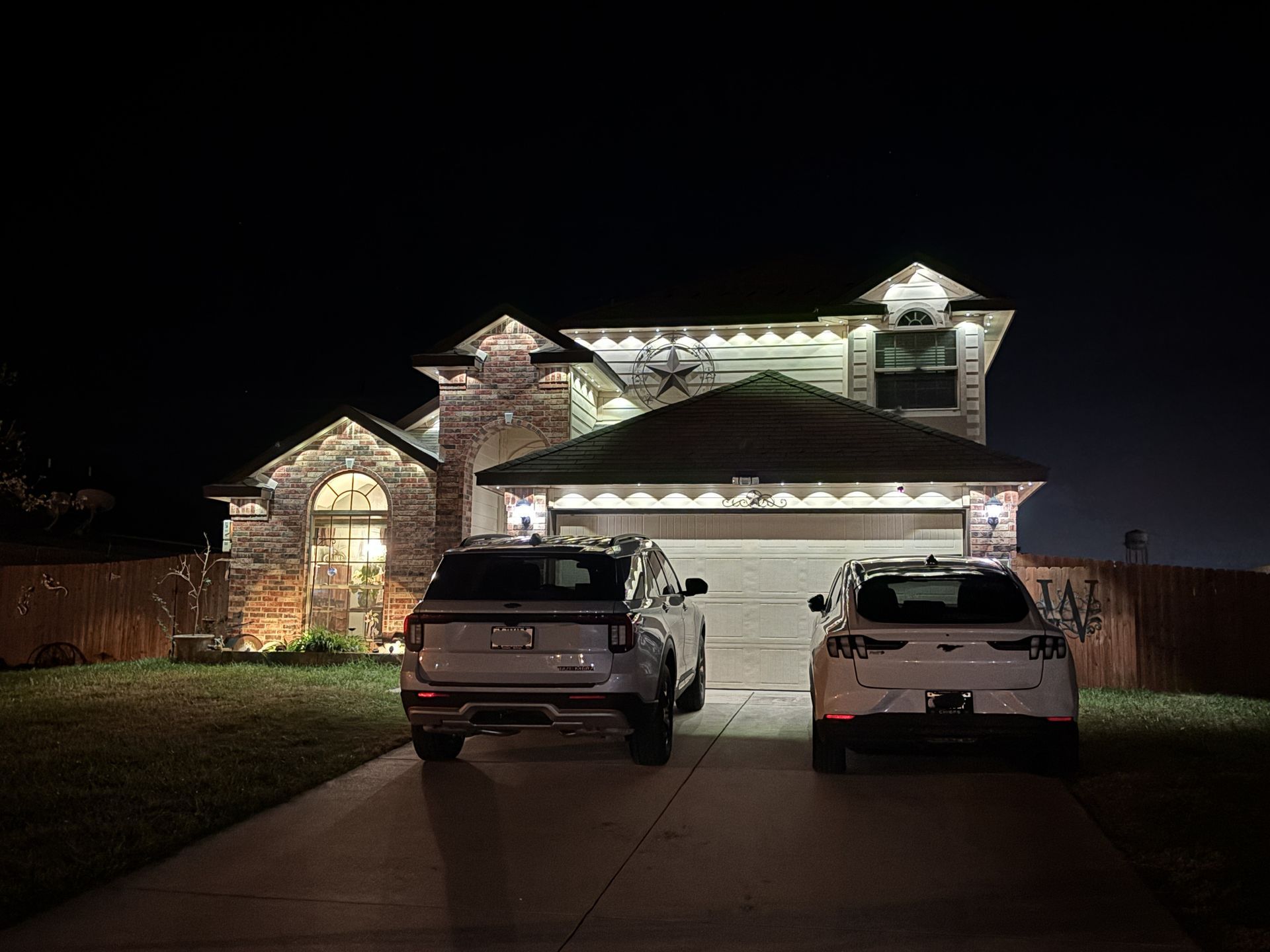 Two white SUVs parked in a driveway in front of a lit house at night