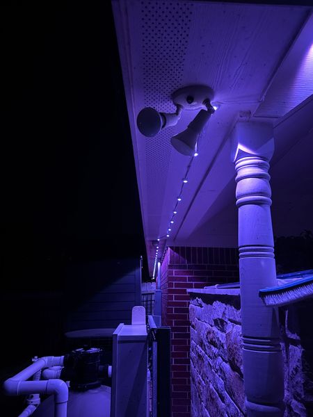 Purple-lit porch ceiling with string lights, a wall-mounted fan, and visible pipes at night