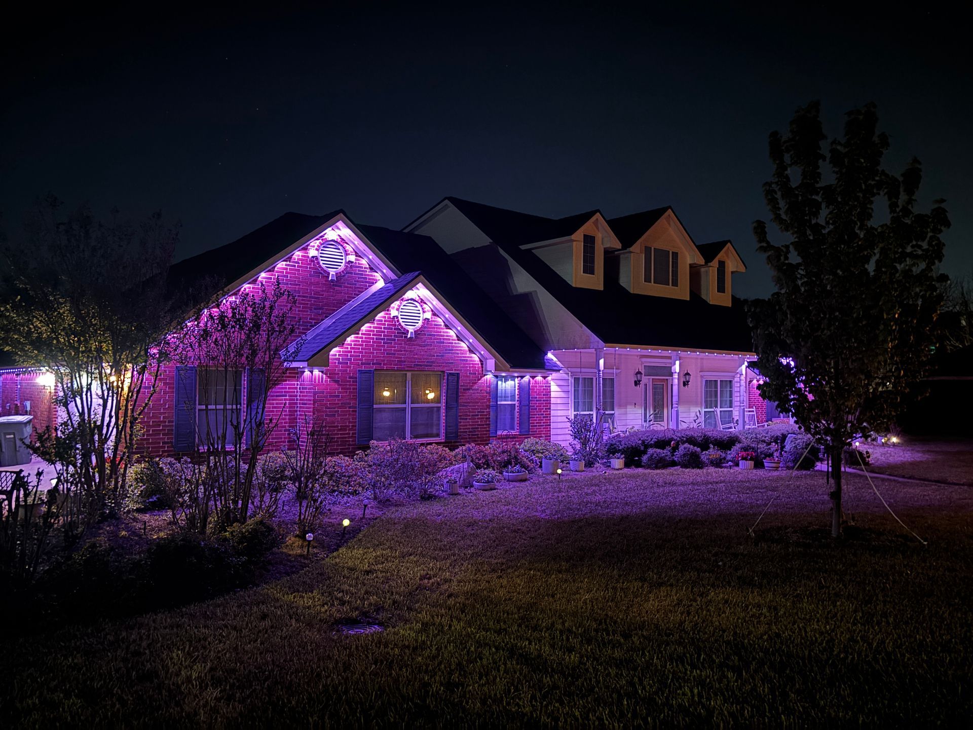House lit with purple holiday lights at night, with snow-covered yard and trees.