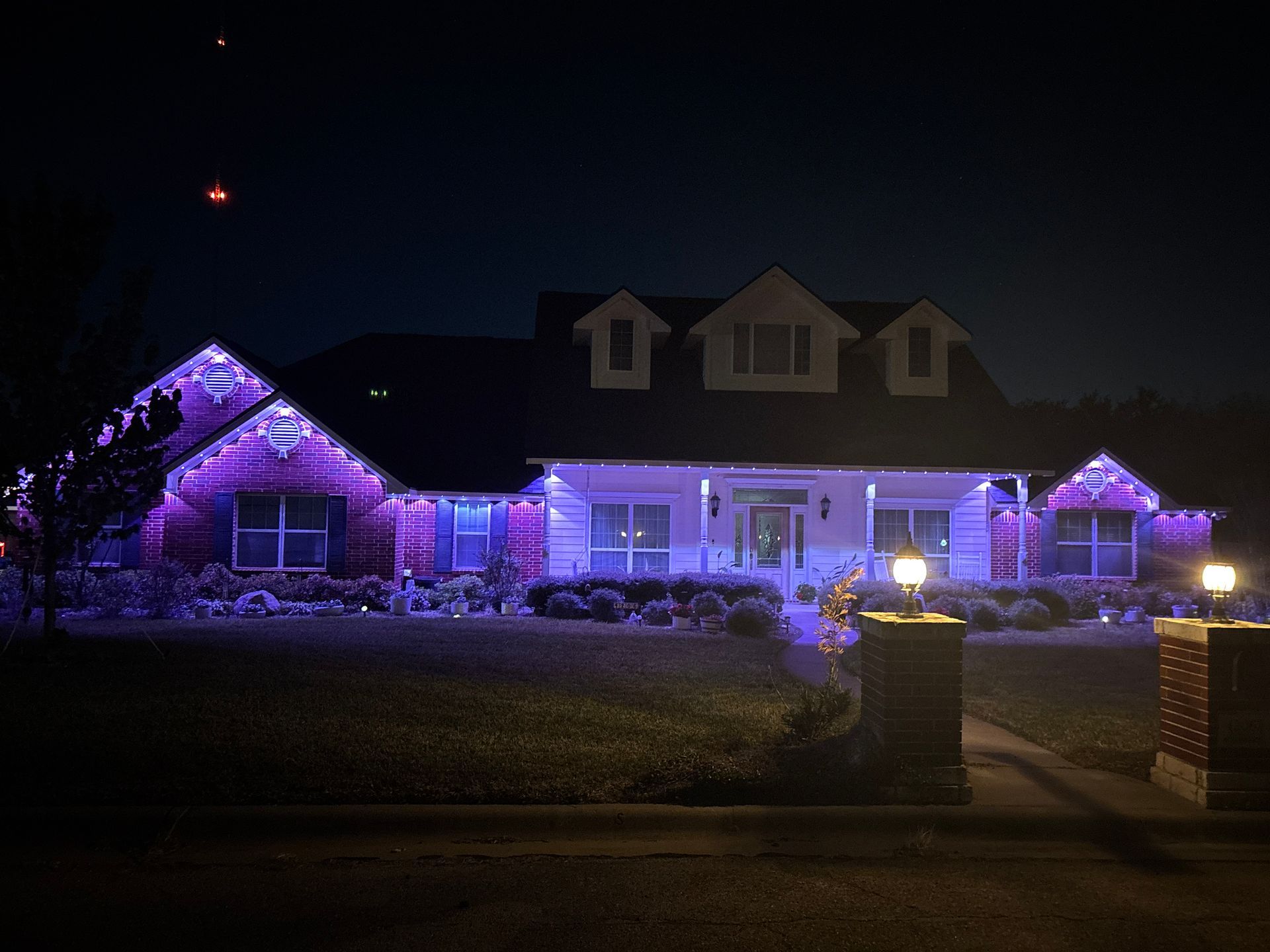 House lit with purple holiday lights at night, viewed from the street behind a small front yard and gate
