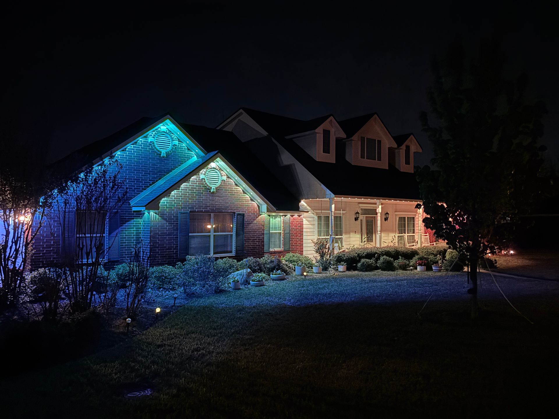 Suburban house at night lit with blue and warm holiday lights, snow on the roof and yard