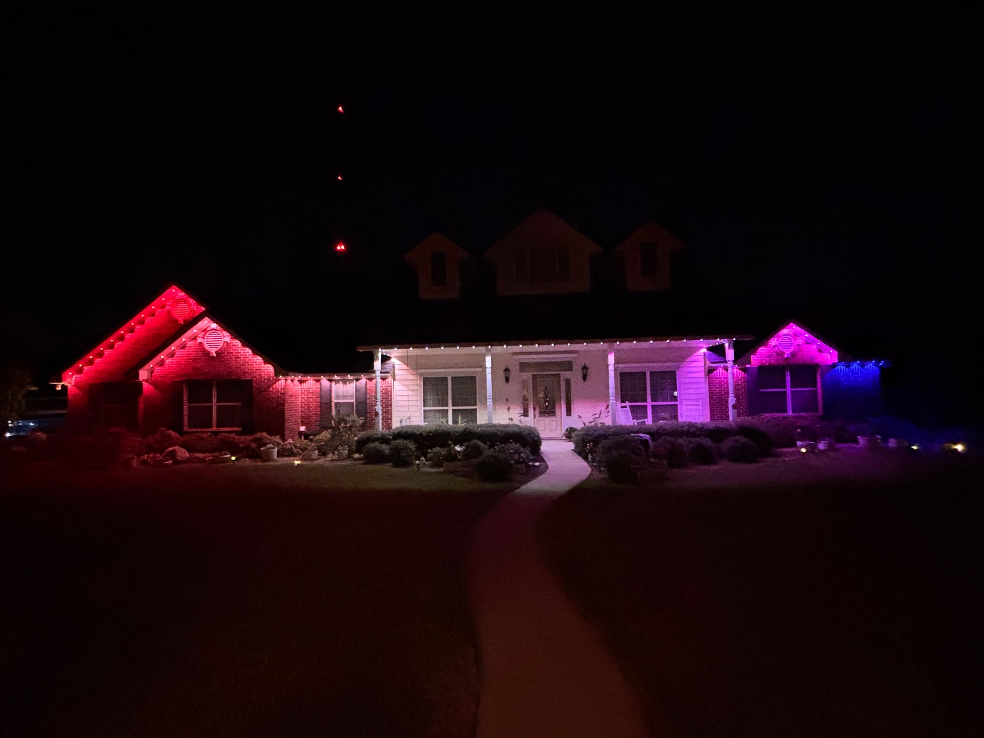 House lit in red, white, and blue holiday lights at night with snow-covered yard and parked cars