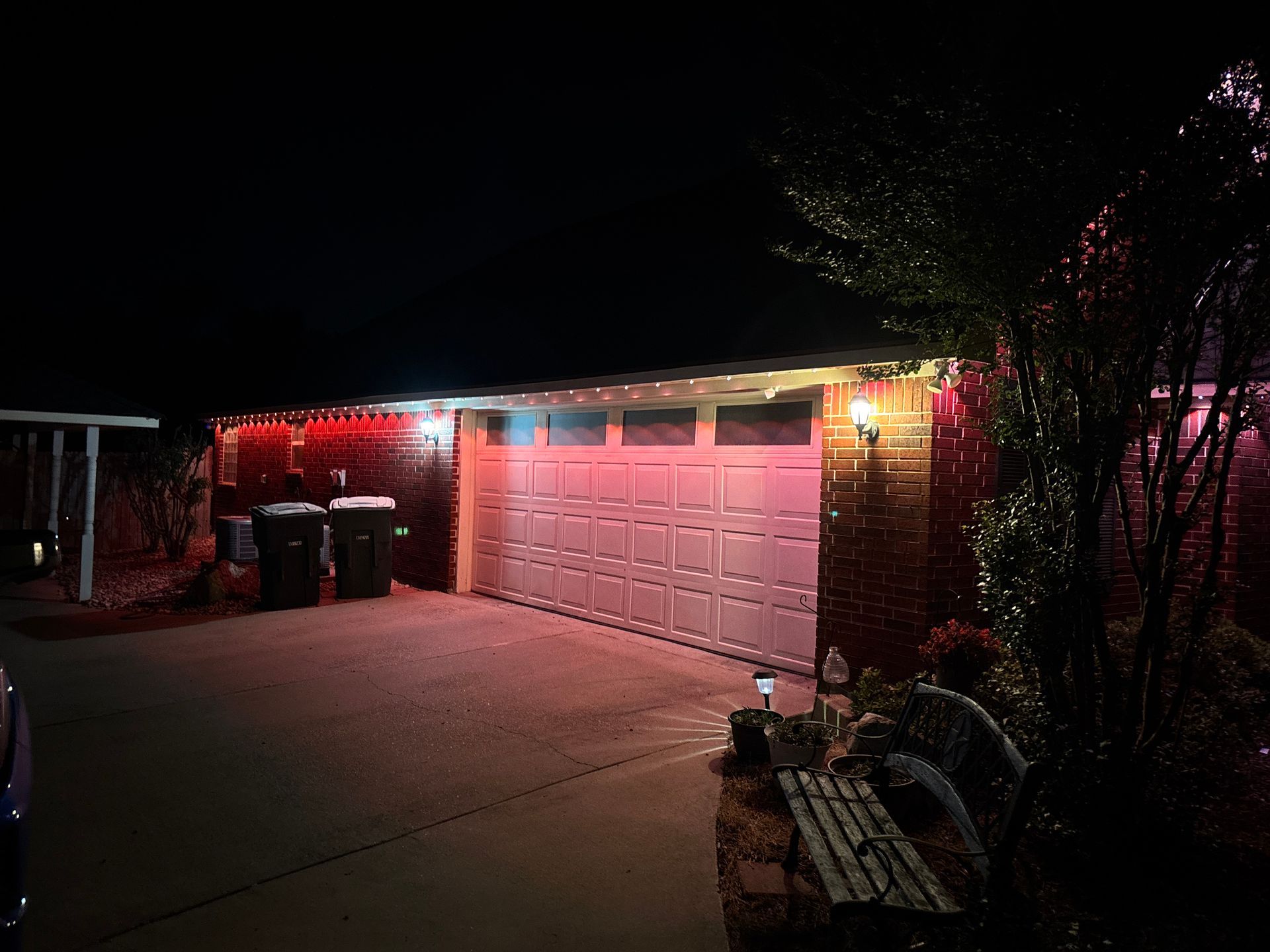 House garage lit by red and pink holiday lights at night, with trash bins and a driveway
