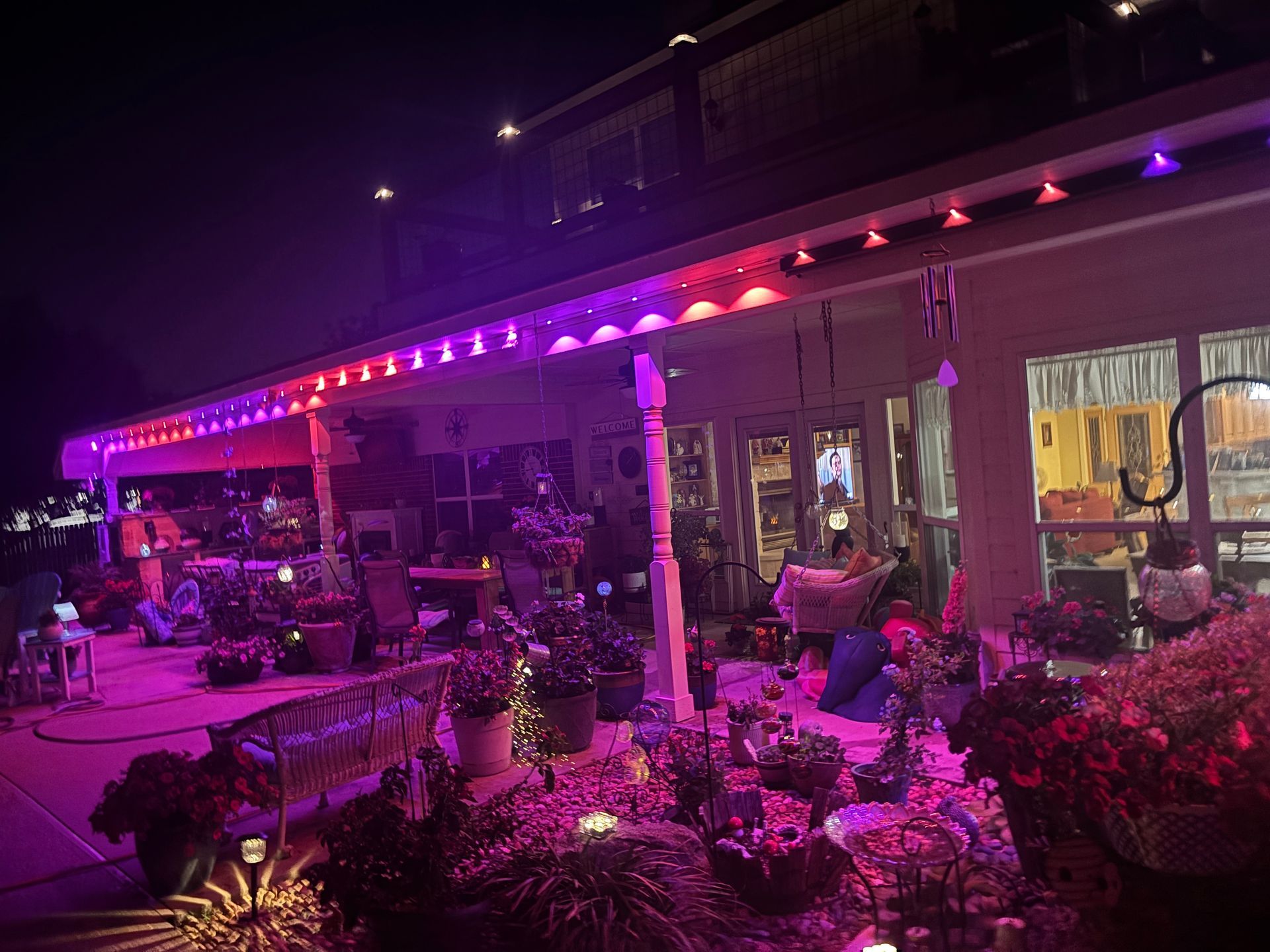 Outdoor patio lit in pink and purple at night, with tables, umbrellas, and potted plants outside a building