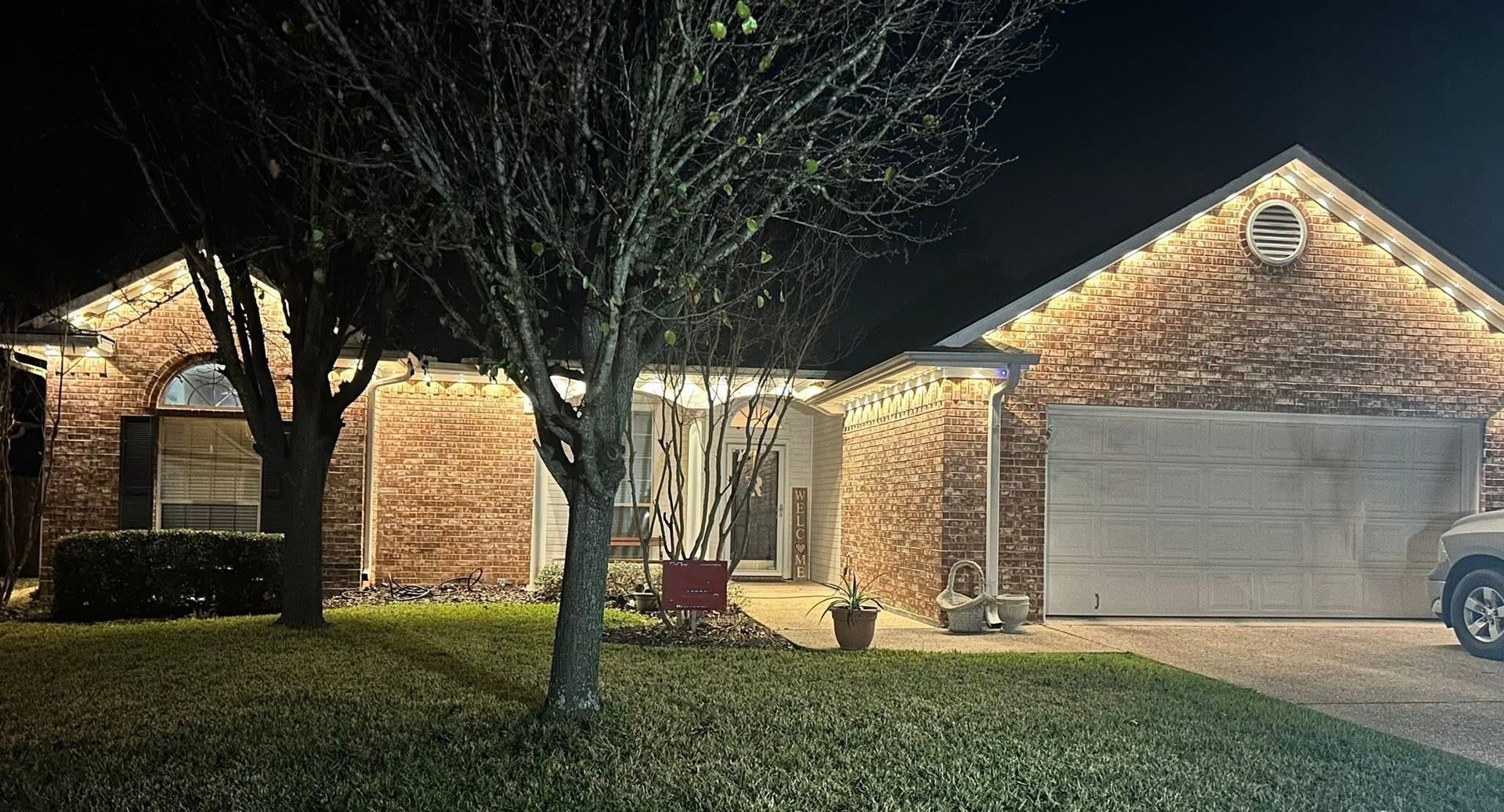 Nighttime view of a brick house with lit exterior lights, a garage, and a tree in the front yard.