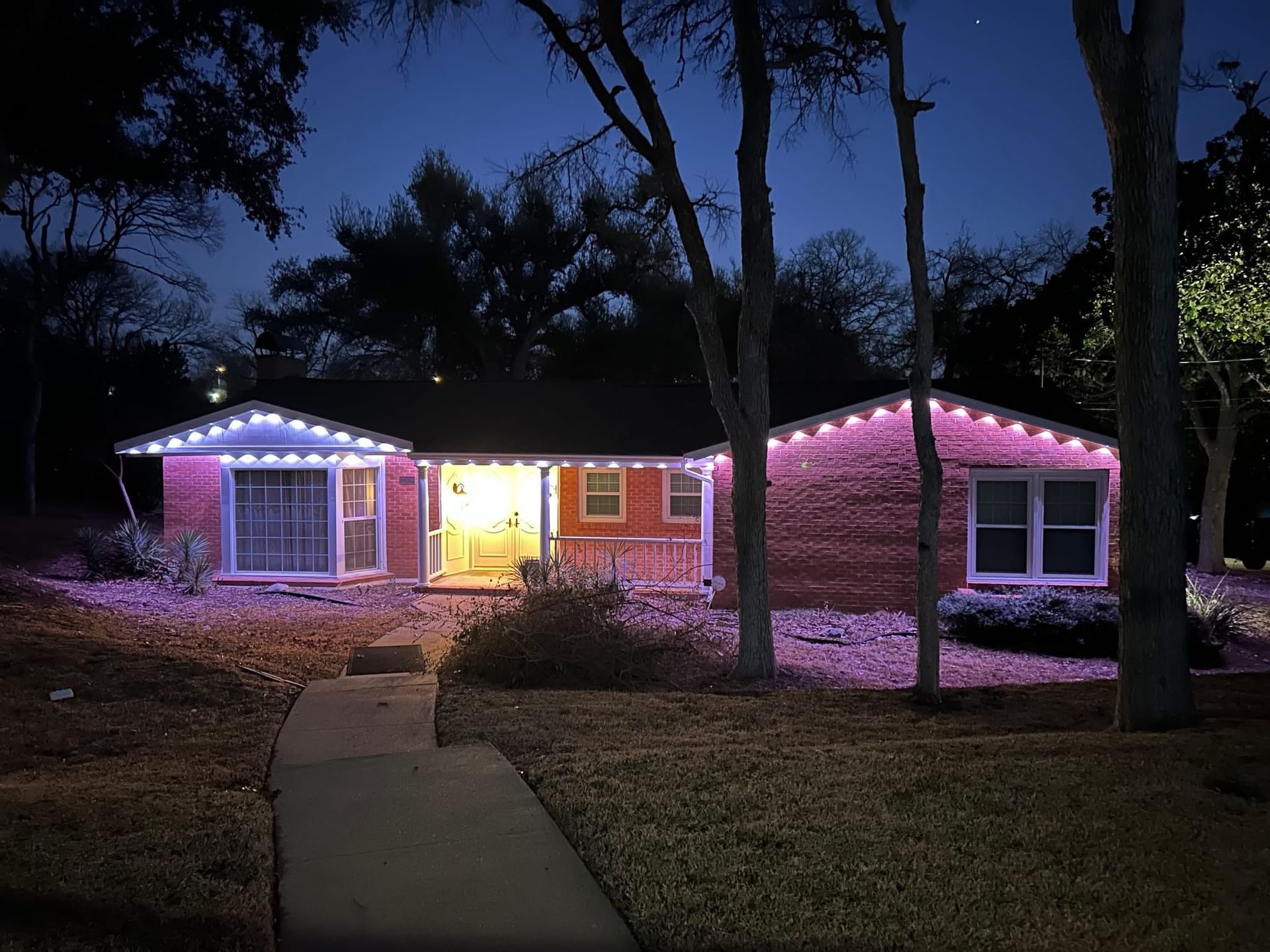 Lit house at dusk with pink and white roof lights, viewed from a sidewalk in a wooded yard.