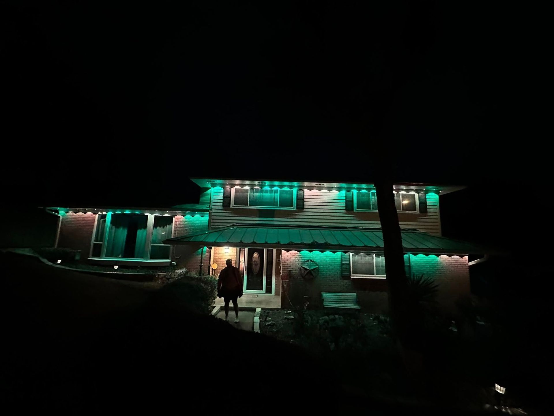 Two-story house lit with green lights at night, with a person standing by the front door.