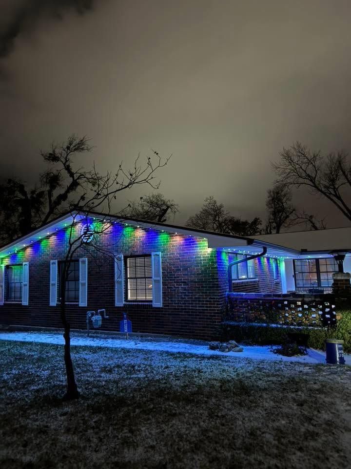 Brick house at night with colorful holiday lights and blue-lit snowy yard under a cloudy sky