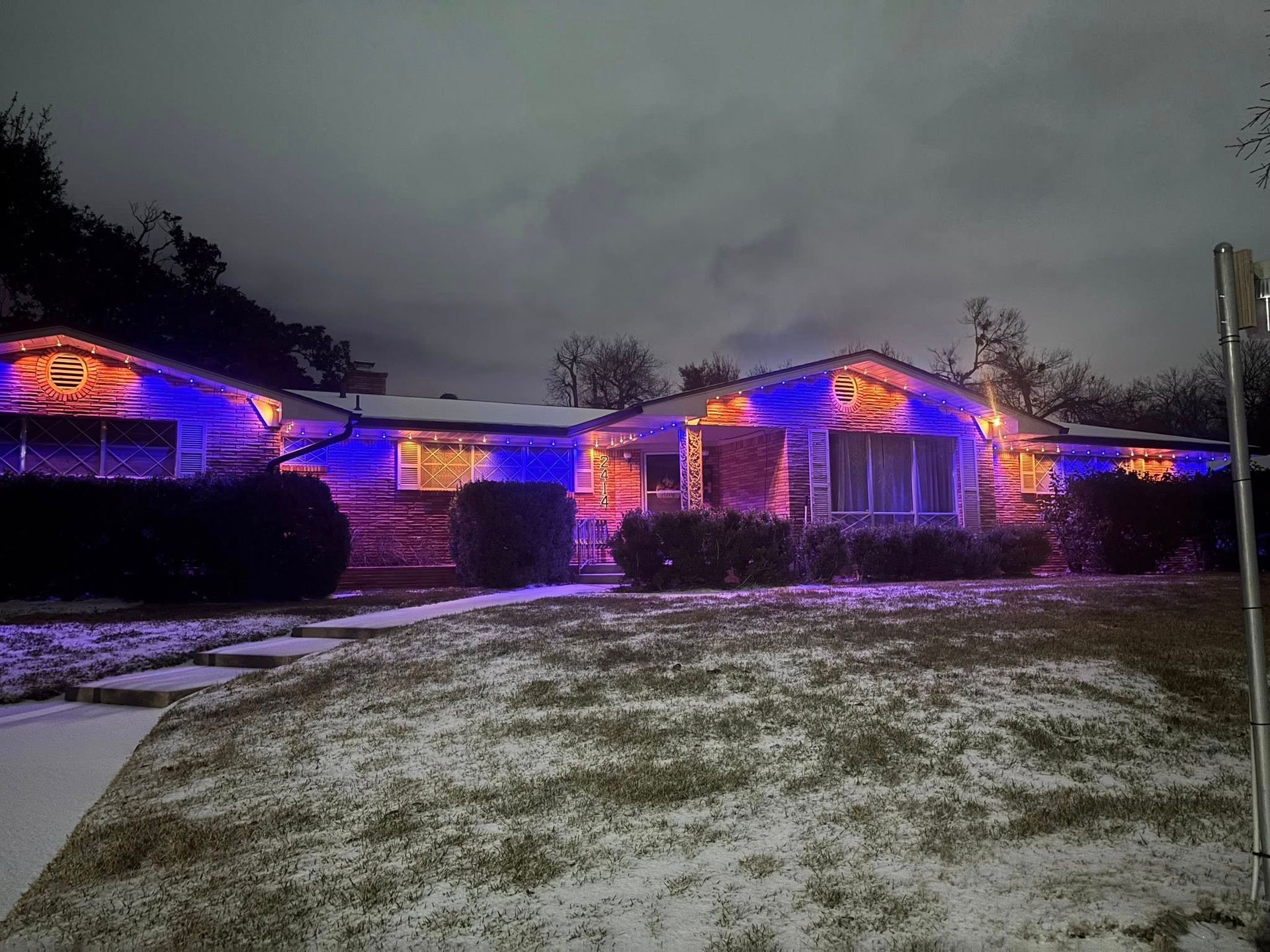 Single-story house lit with purple and orange lights at dusk, with a snowy front yard.