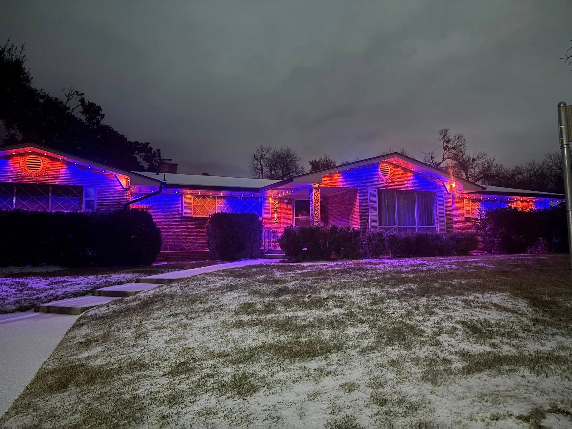 Single-story house at dusk lit with red and blue holiday lights, with light snow on the lawn.