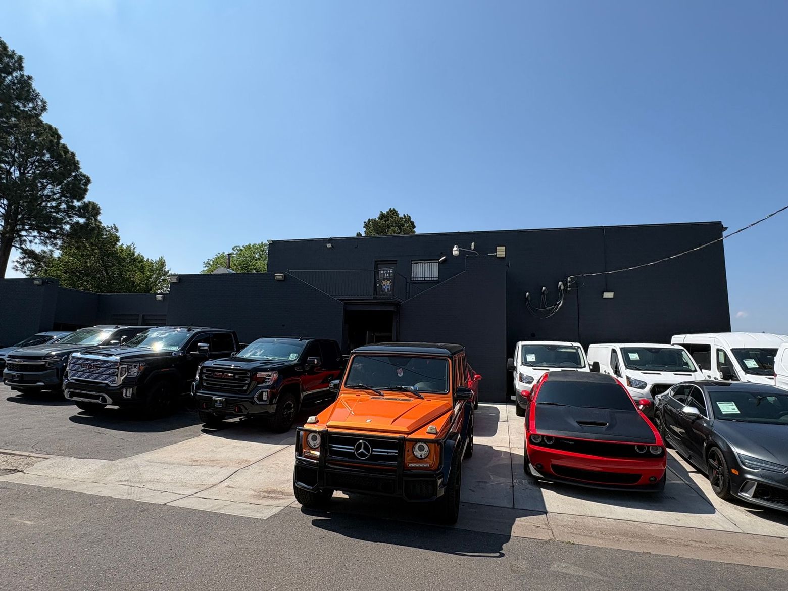 Parked cars in front of a black building, including an orange SUV and a red sedan under a clear blue sky