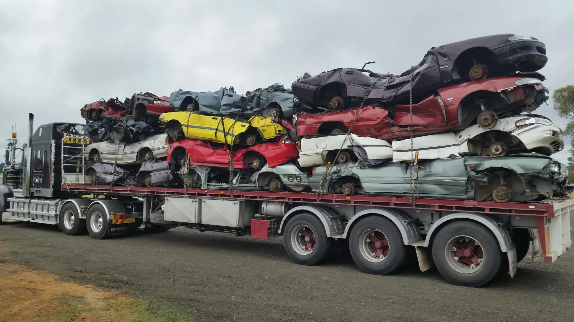 Car carrier truck loaded with stacked wrecked cars in white, red, black, and yellow on a road.