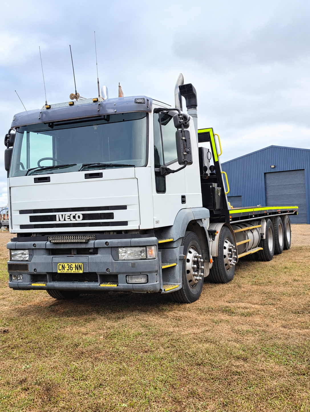 White Iveco tow truck with flatbed parked on grass beside a blue industrial building under cloudy sky