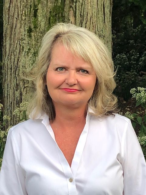 A woman in a white shirt is standing in front of a tree.