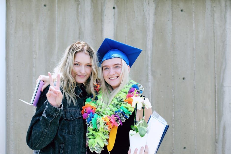 Two people smiling, one in a blue graduation cap with a rainbow lei, standing against a concrete wall.