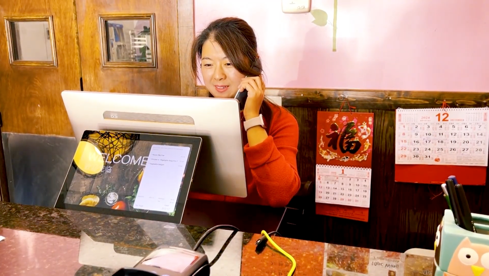 A Chinese, Asian woman, who appears to be the restaurant owner, is standing behind the checkout counter using a ZBS POS (point of sale) system Front Station. She is on the phone, entering an order while managing the front station.