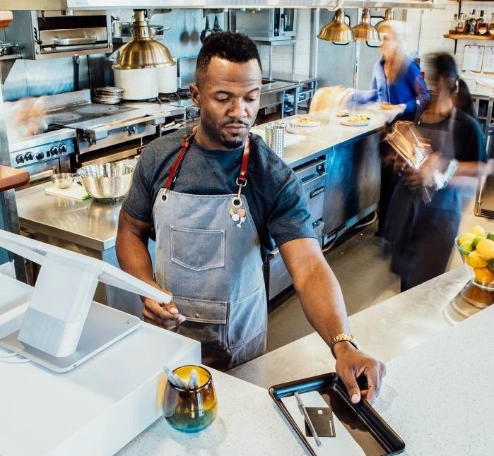 Male employee standing behind the cashier counter at an open-concept restaurant, handing a check to a customer. The restaurant features a modern design with a visible open kitchen area in the background, and the cashier counter includes a Clover POS system for transactions