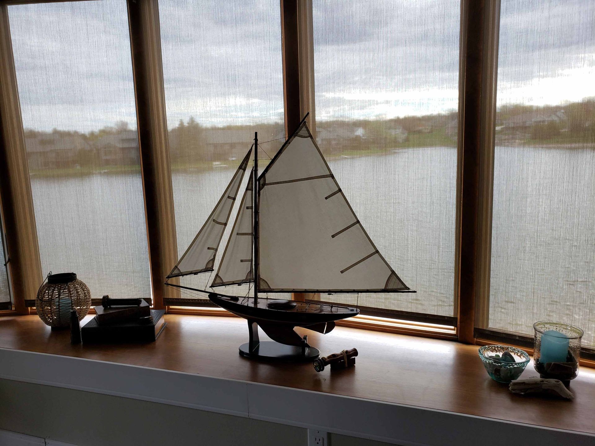 A model sailboat sits on a window sill in front of a window overlooking a body of water.
