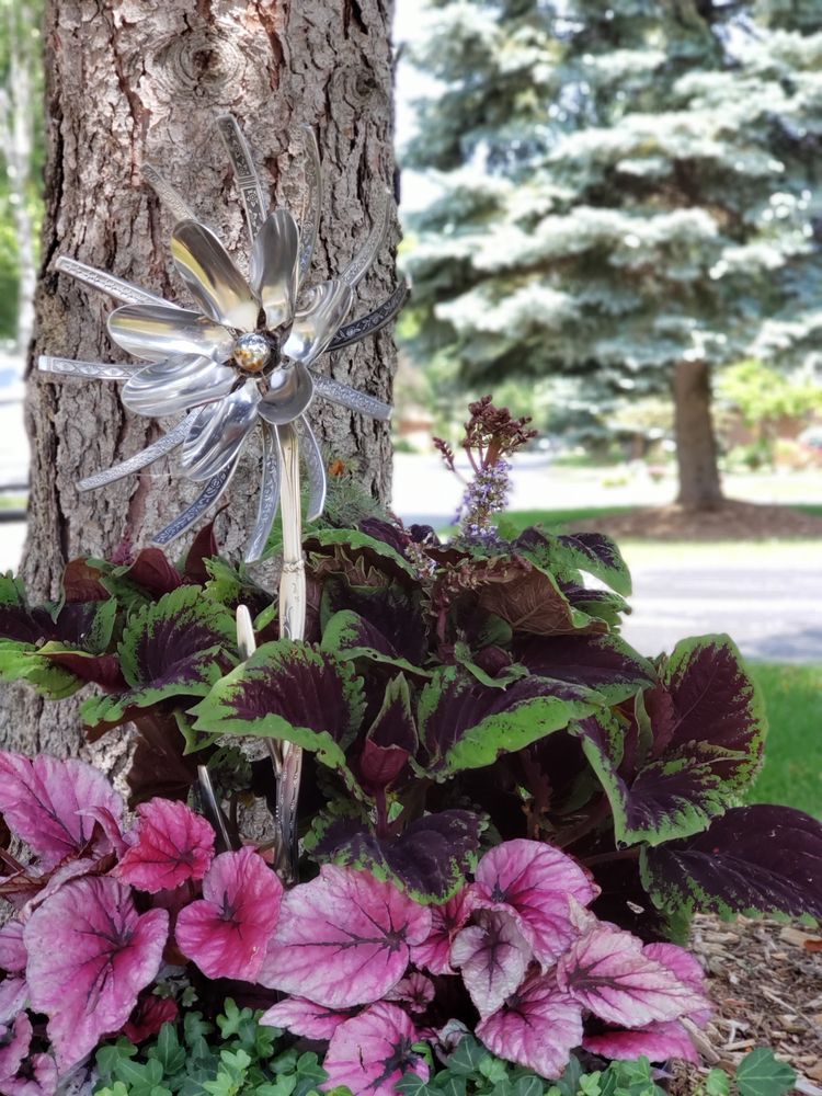 A windmill made out of spoons is sitting next to a tree in a garden.