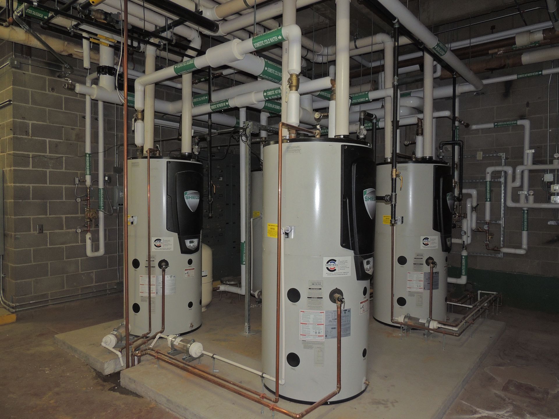 Three water heaters are lined up in a room with pipes