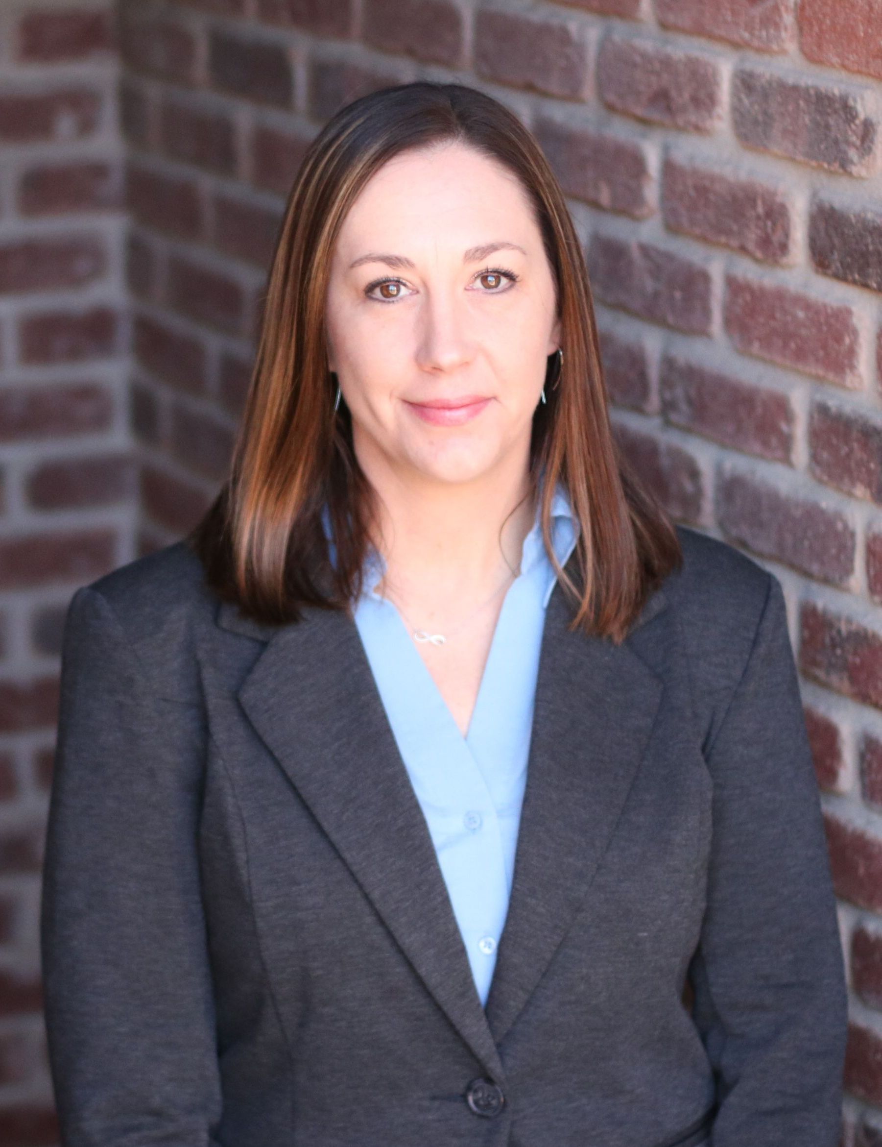 A woman in a suit and blue shirt is standing in front of a brick wall.
