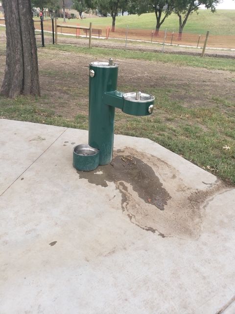 A green water fountain is sitting on a sidewalk in a park