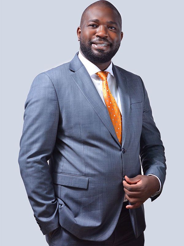 Man in grey suit and orange tie smiles, standing against a light grey backdrop.
