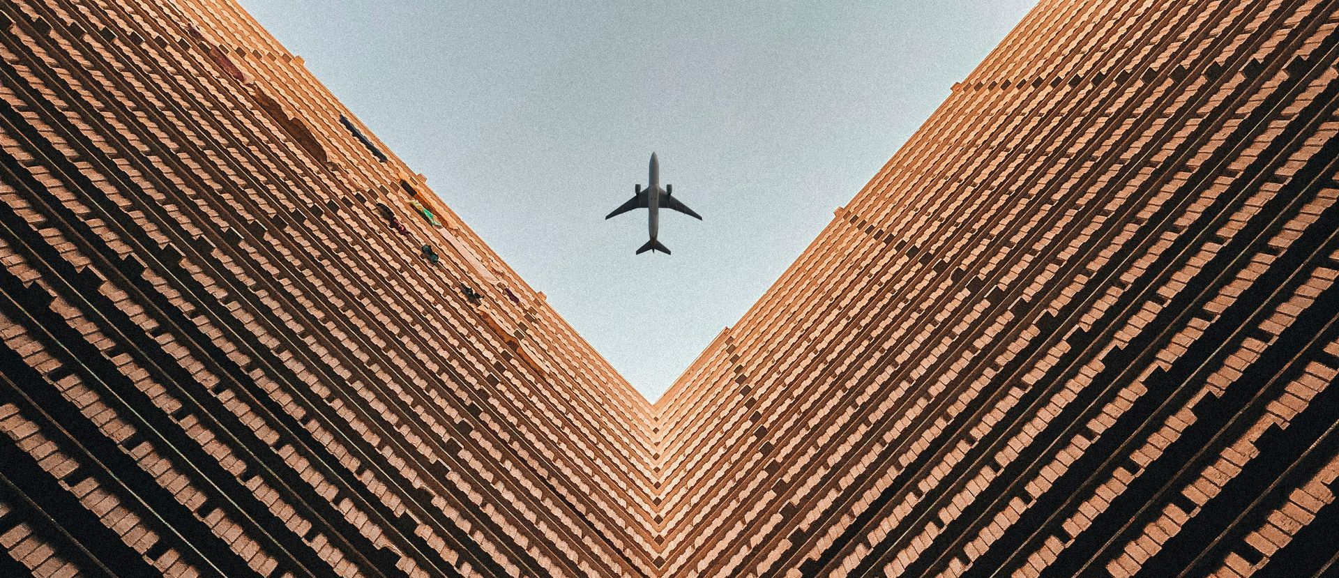 A jet flies through the sky framed by two tall buildings with a brown facade.