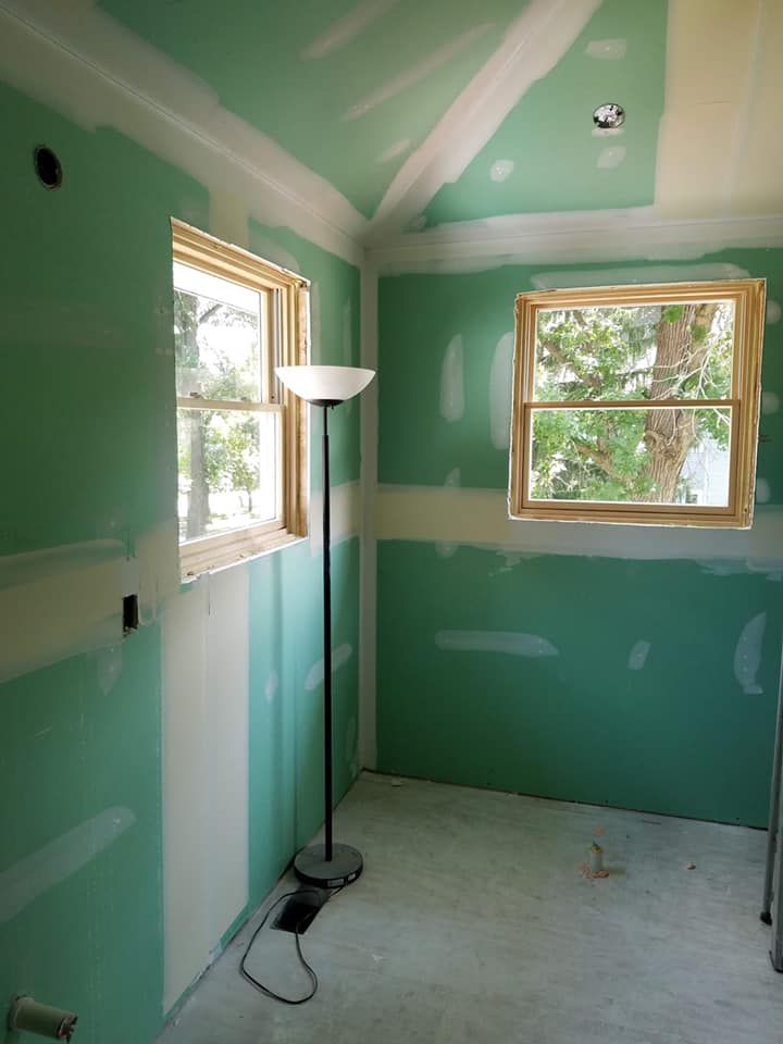 Green room under renovation with white drywall patches, two windows, and a floor lamp.