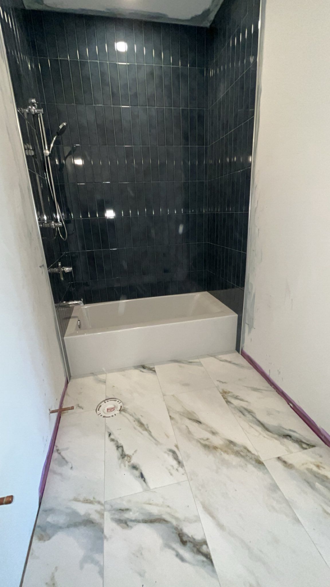 Narrow tiled bathroom alcove with dark wall tile, white tub base, and marble-look floor tiles.