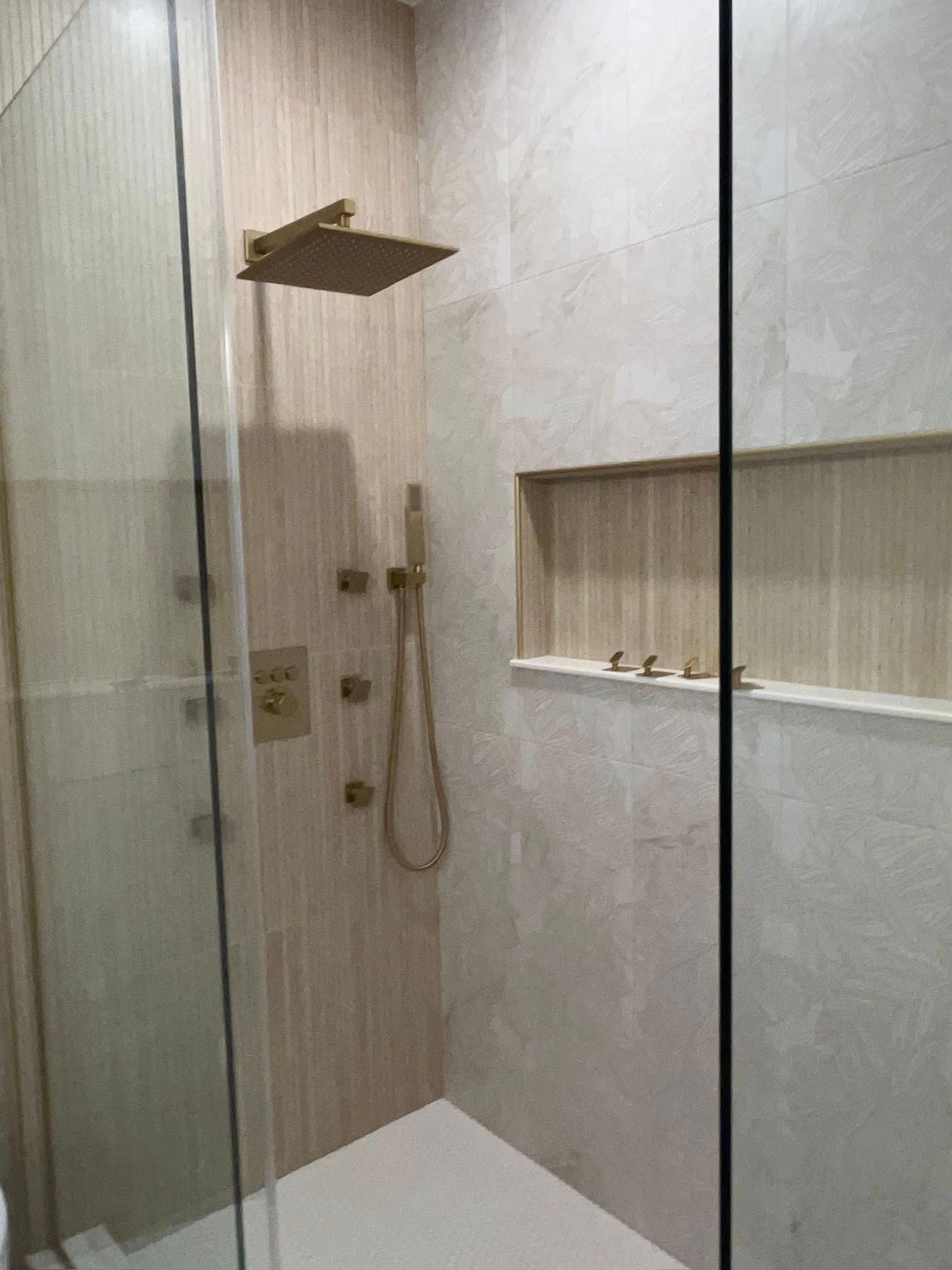 Glass shower stall with rain shower head, handheld sprayer, and built-in shelf in beige tiled bathroom