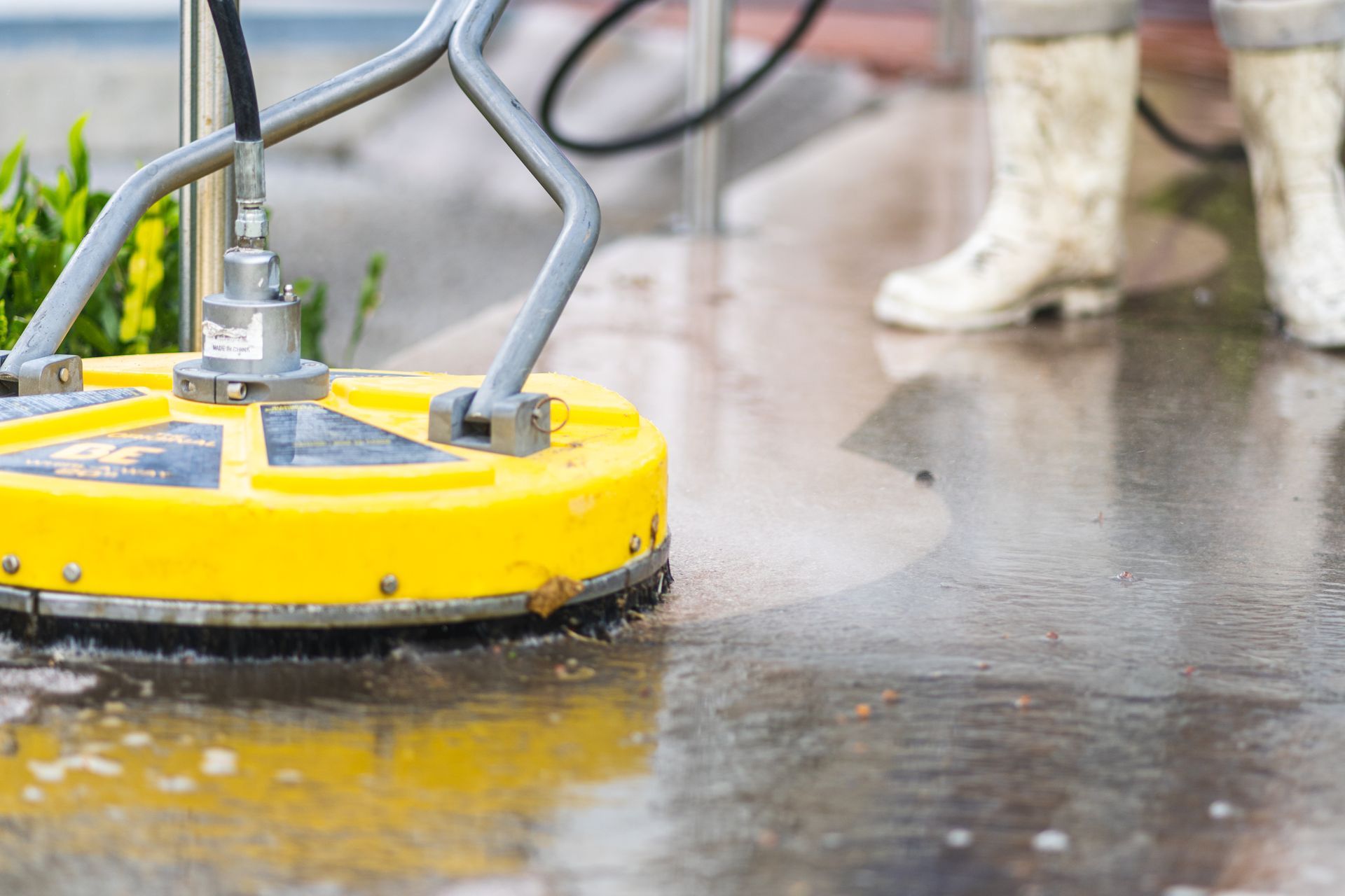 Yellow surface cleaner on a concrete surface, spraying water. A person in white boots stands nearby.