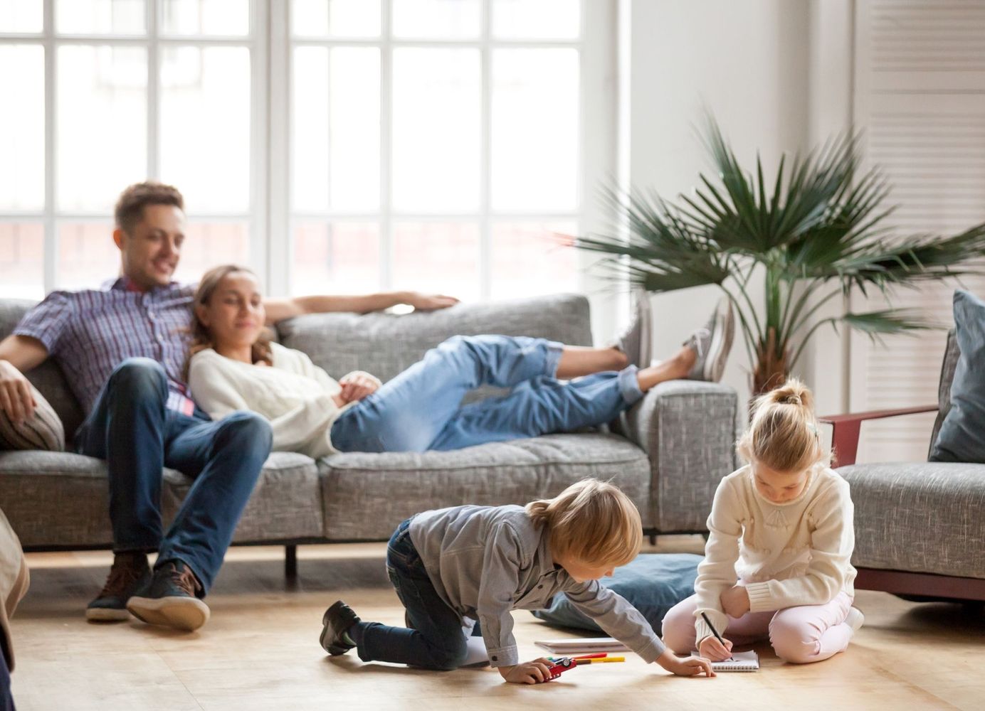 Family relaxing on a gray sofa while two children play on the wooden floor in a bright living room