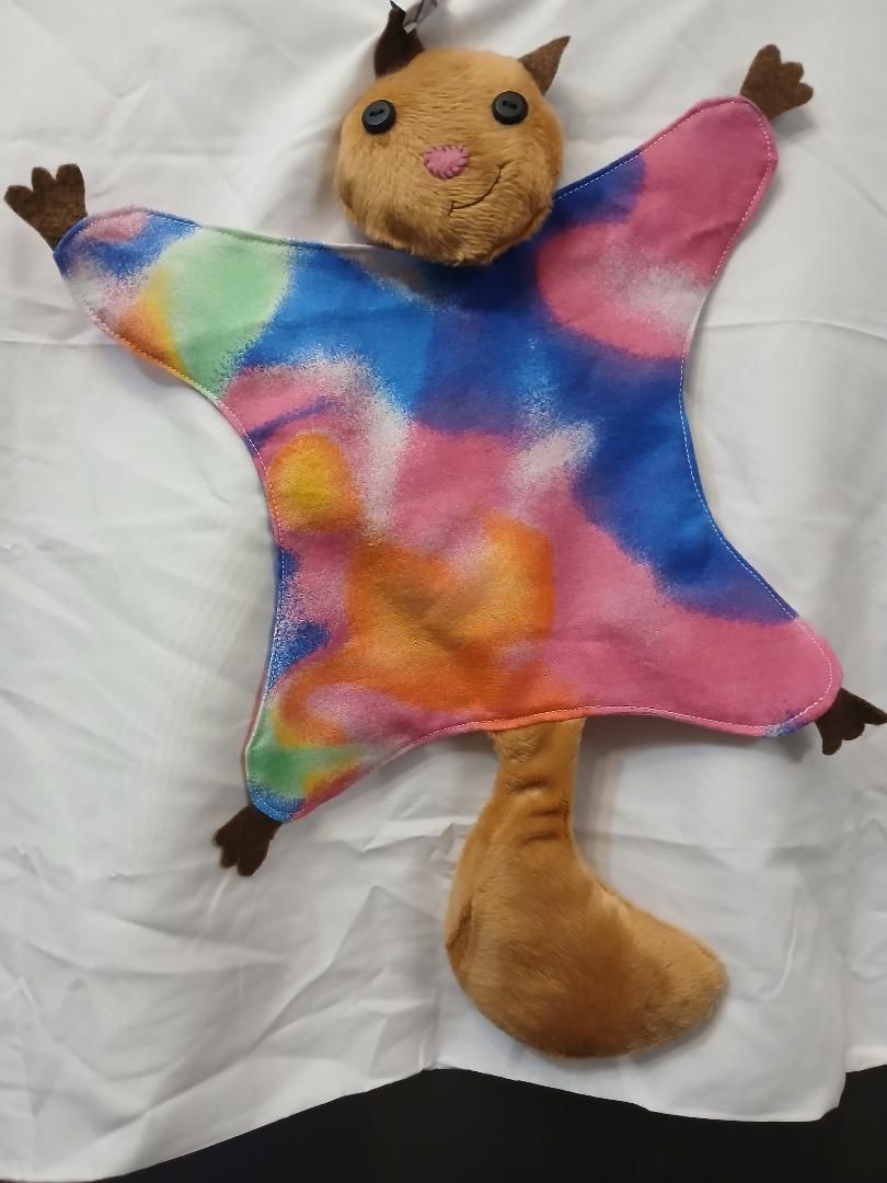 Colorful plush toy with a tan face and limbs, wearing a tie-dye body on a white background
