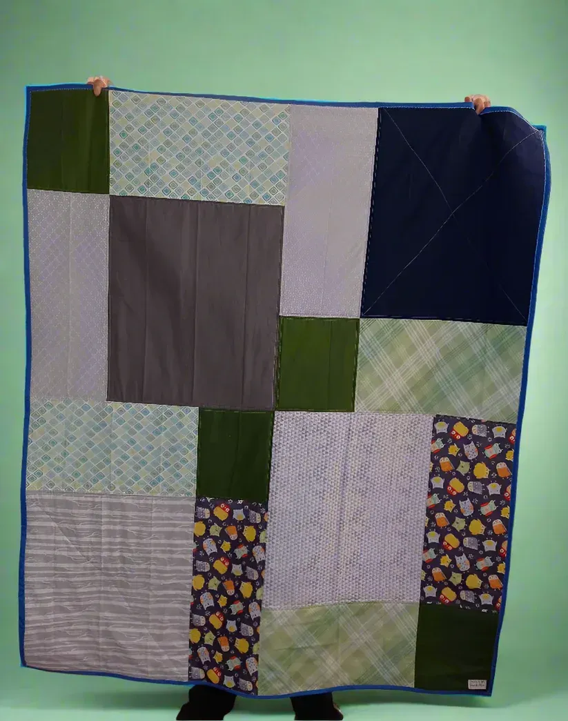 Patchwork quilt in blue, green, gray, and white squares hanging against a plain background