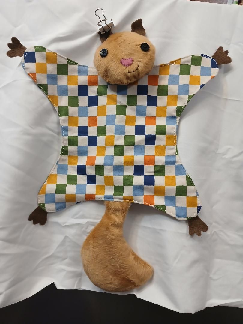 Handmade cat-shaped fabric toy with checkered patchwork body, tan face and tail, and tiny paw tassels on a white background