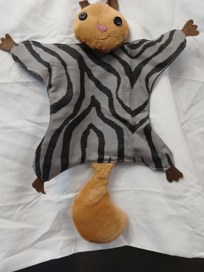 Plush cat toy with gray striped body, tan head and tail, lying on white bedding with arms outstretched