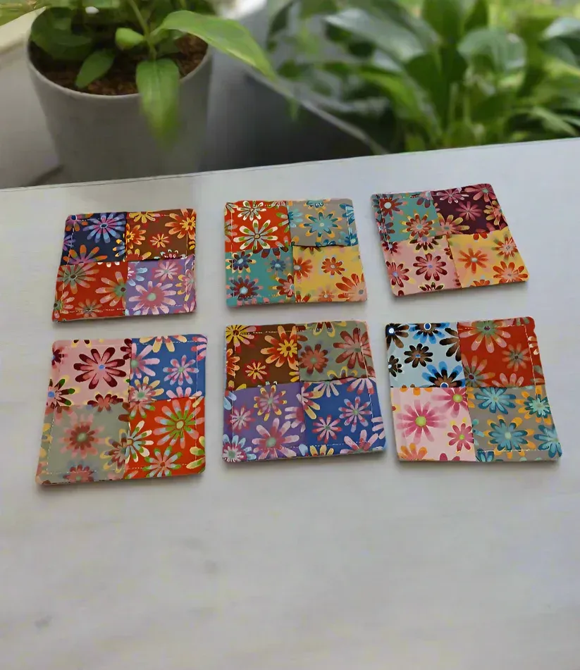 Six colorful floral square coasters on a table, with potted plants blurred in the background