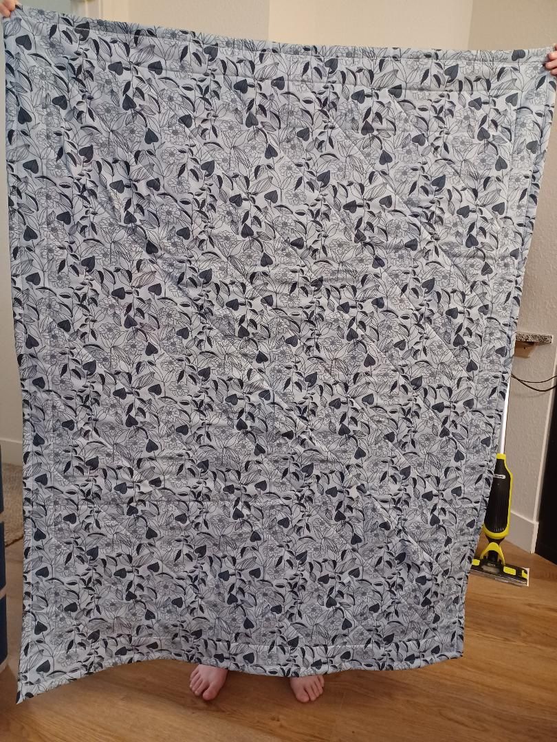 Person standing behind a large gray-and-black patterned blanket indoors, with bare feet visible at the bottom