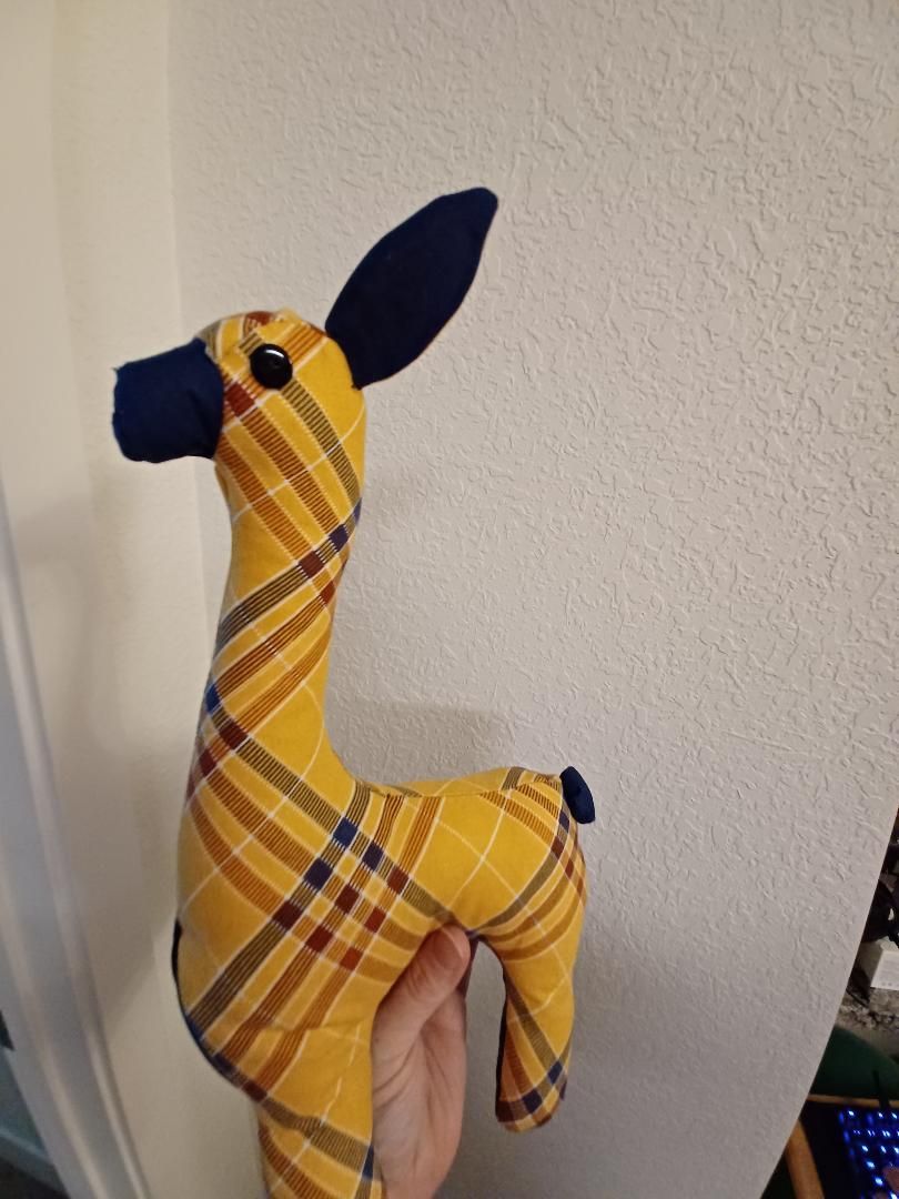 Yellow plaid stuffed giraffe with black ears and snout, held upright against a plain wall.