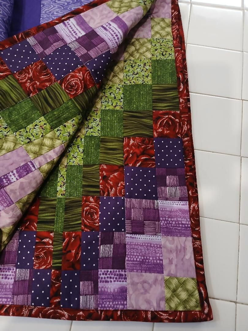 Patchwork quilt with red, green, purple, and pink squares draped over a white tiled wall.