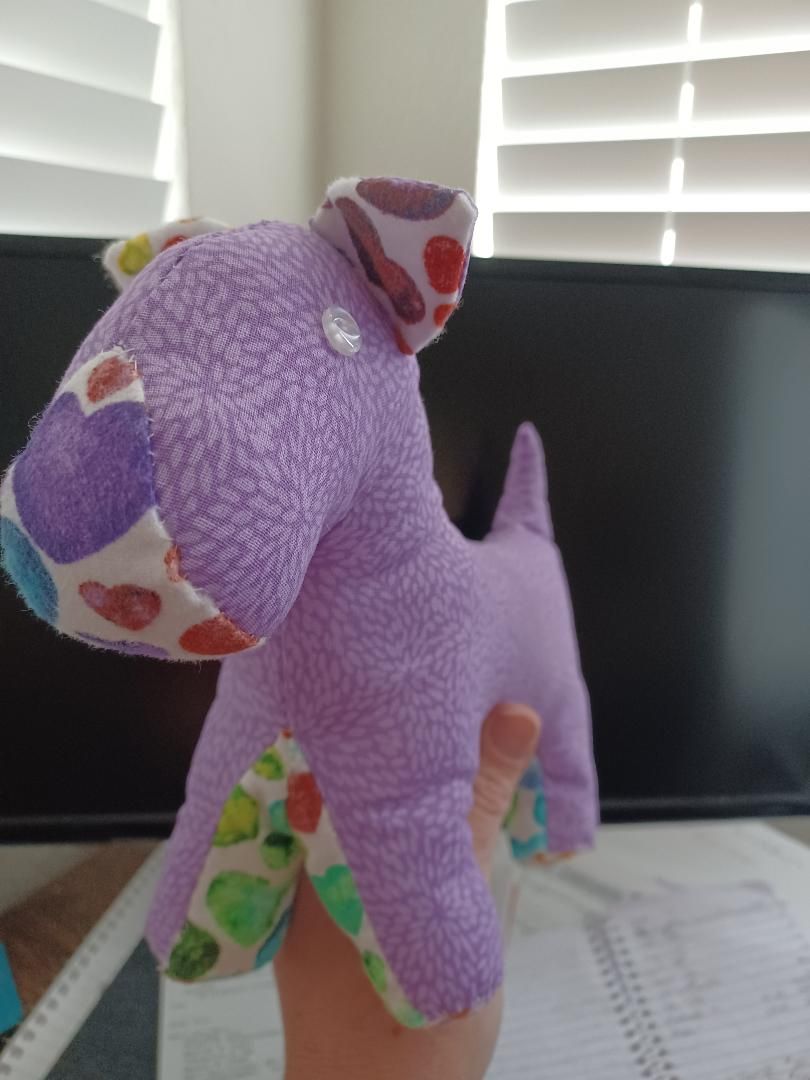 Purple plush dinosaur toy with colorful polka dots standing on a table by window blinds