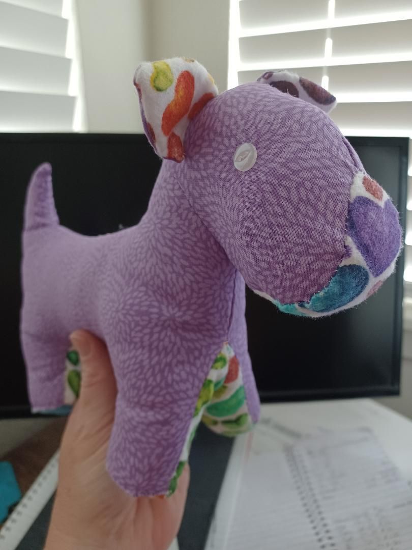 Purple plush dog toy with colorful patches, held in a hand in front of a computer monitor and window blinds