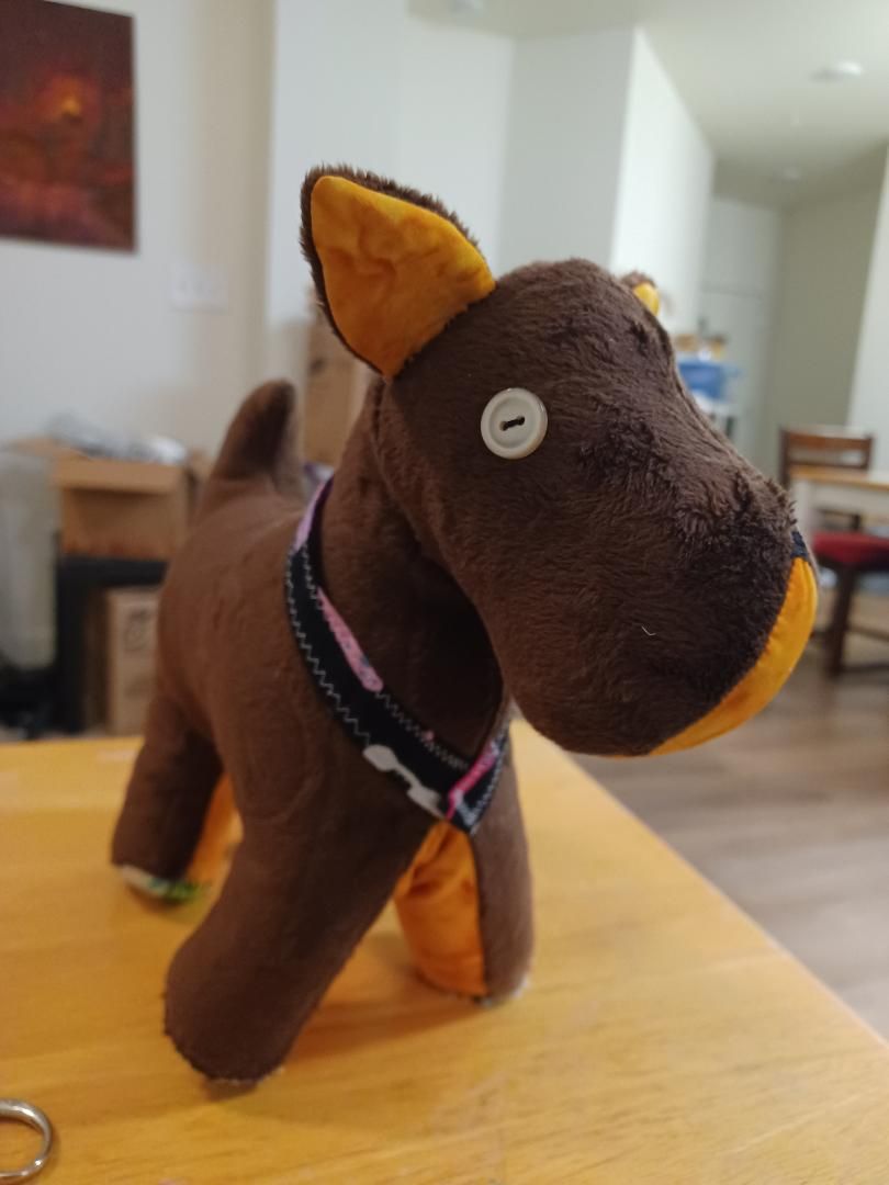 Small brown stuffed horse toy with white button eye and pink collar standing on a wooden table in a room