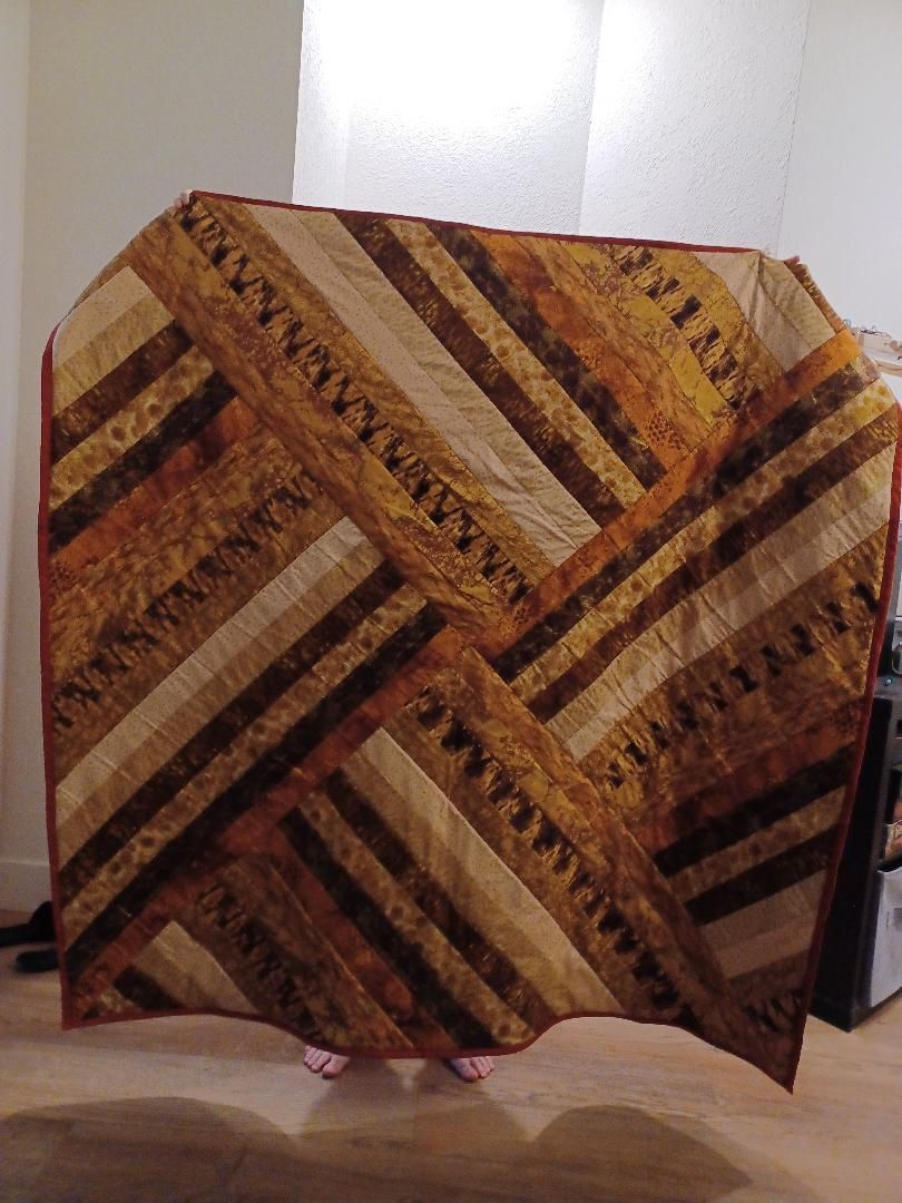 Patterned quilt with tan, brown, and maroon diagonal stripes hanging against a white wall