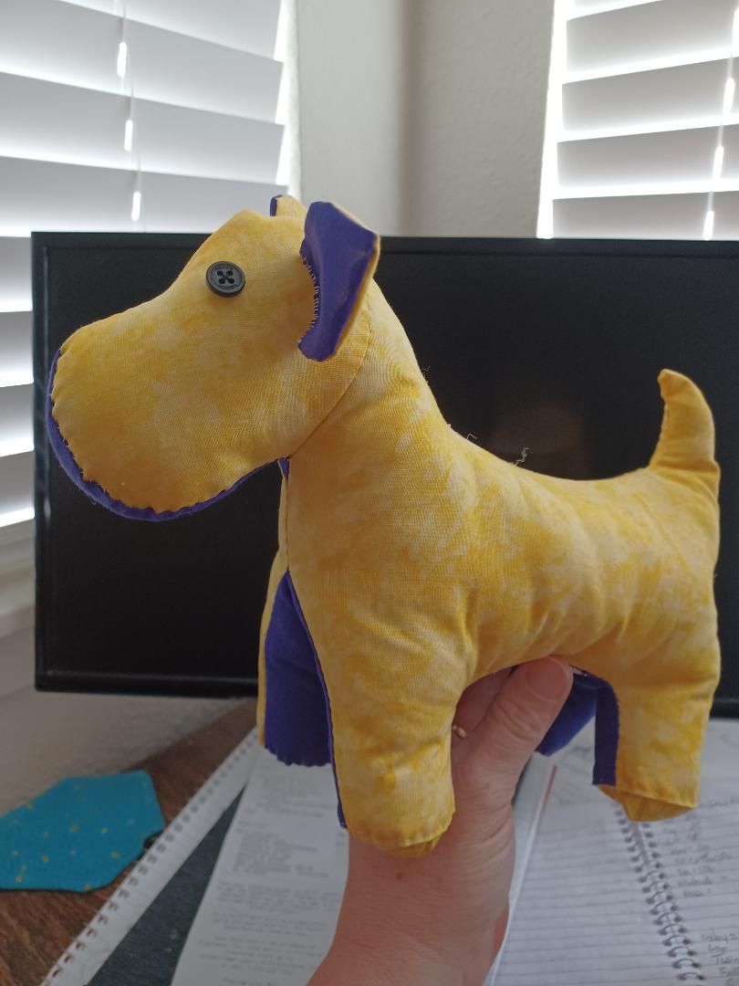 Yellow stuffed dog with purple ears and collar, held up in front of a window with blinds